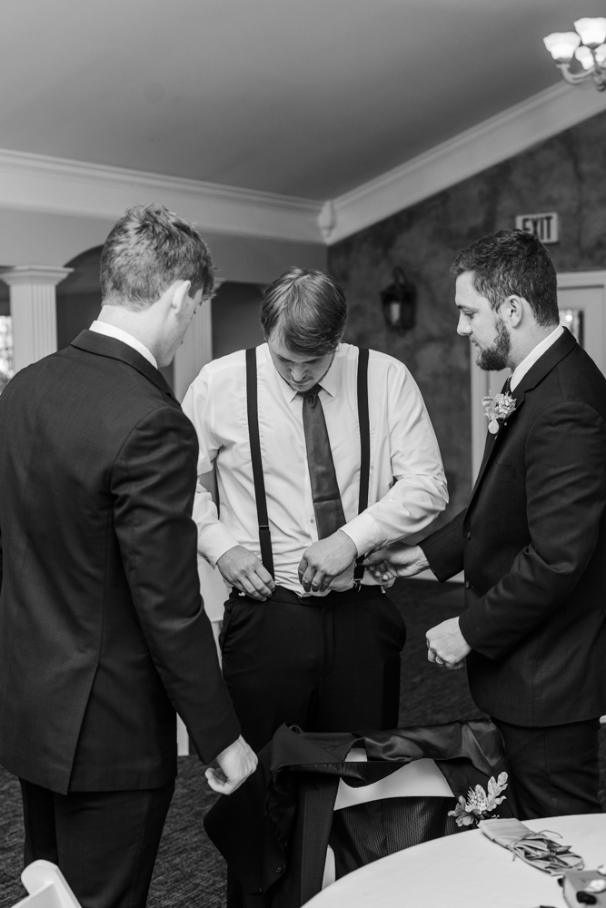 Groom surrounded by his groomsmen as he attaches his suspenders to his suit pants.