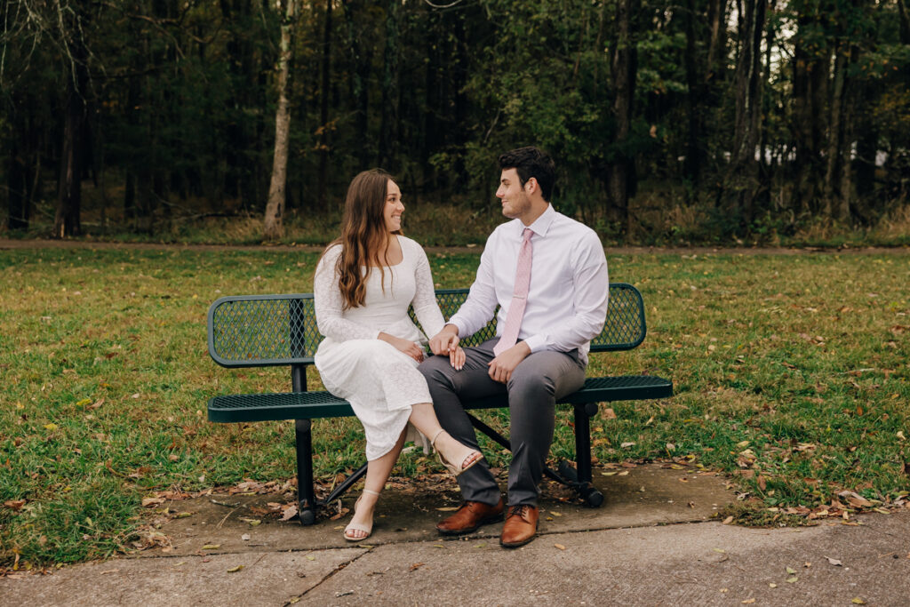 Engaged couple sitting on a green park bench, gazing into each other’s eyes