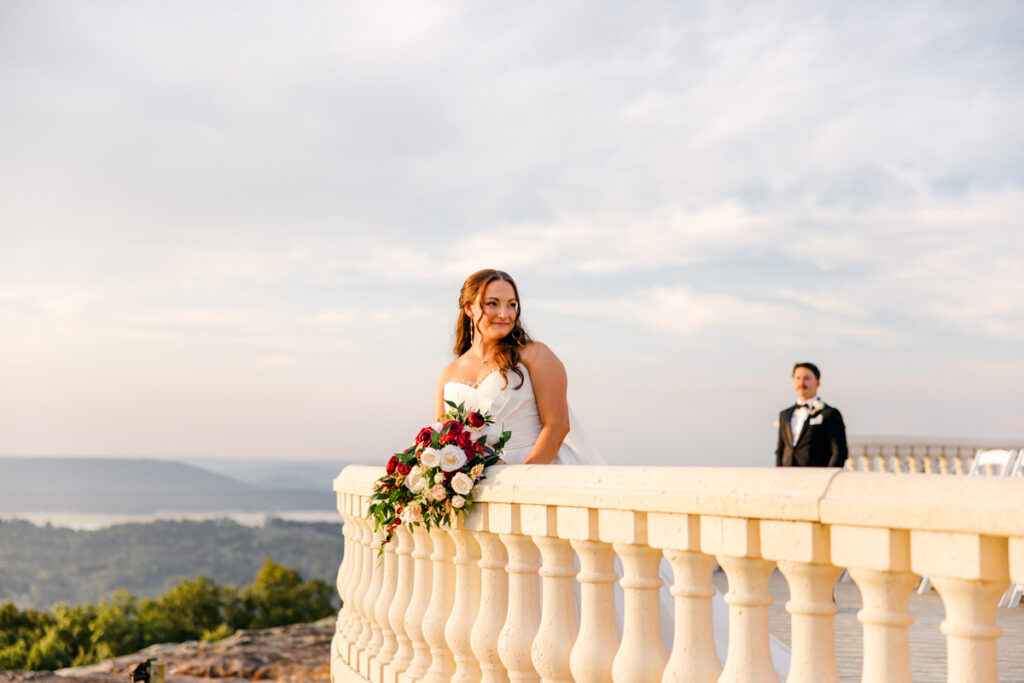 Bride leaning on a railing while her groom admires her from a distance.