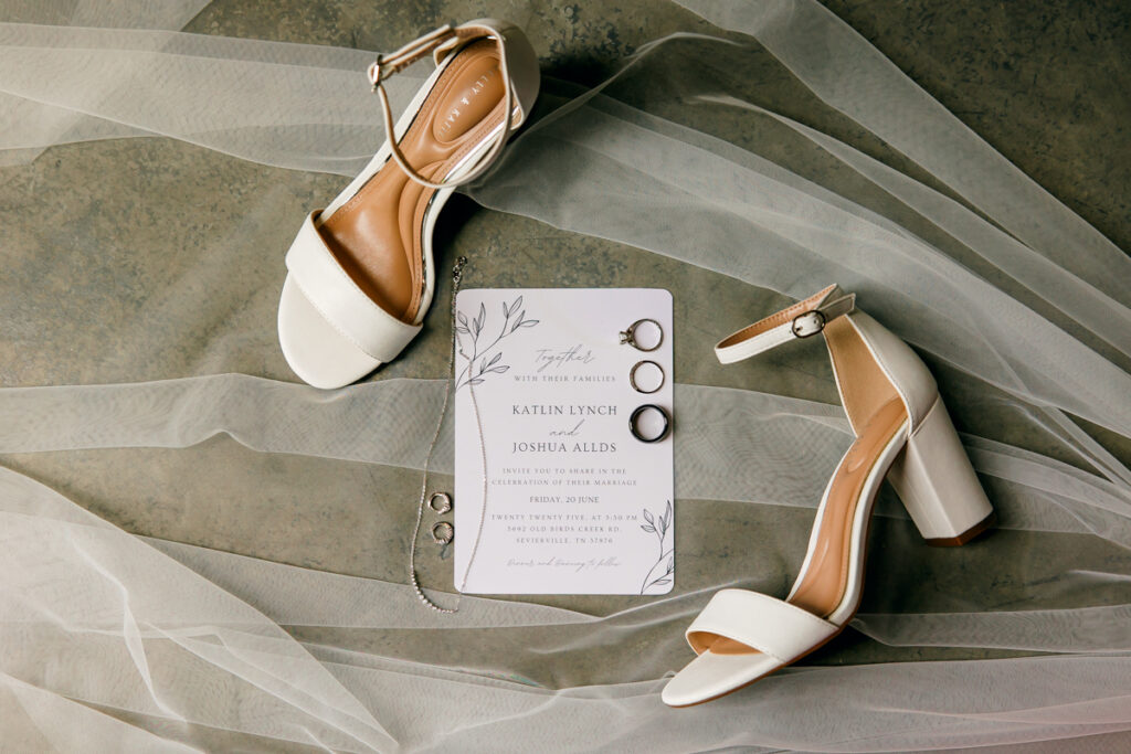 Flat lay of bridal shoes, invitation, necklace, earrings, and rings on a tulle veil