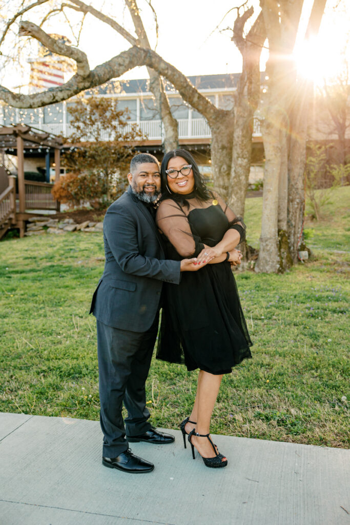 Engaged couple smiling with sunlight streaming through tree branches in the background